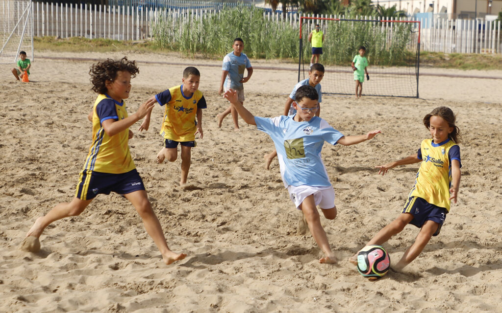 Group of children playing beach soccer on a sandy field, chasing a colorful ball with a yellow and blue jersey on the left and a blue jersey in the center.
