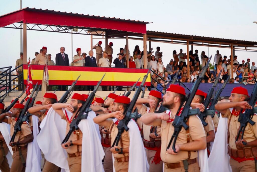Soldiers in khaki uniforms with red berets march in formation, rifles at their shoulders, as spectators watch from bleachers with a large red-yellow Spanish flag banner nearby.