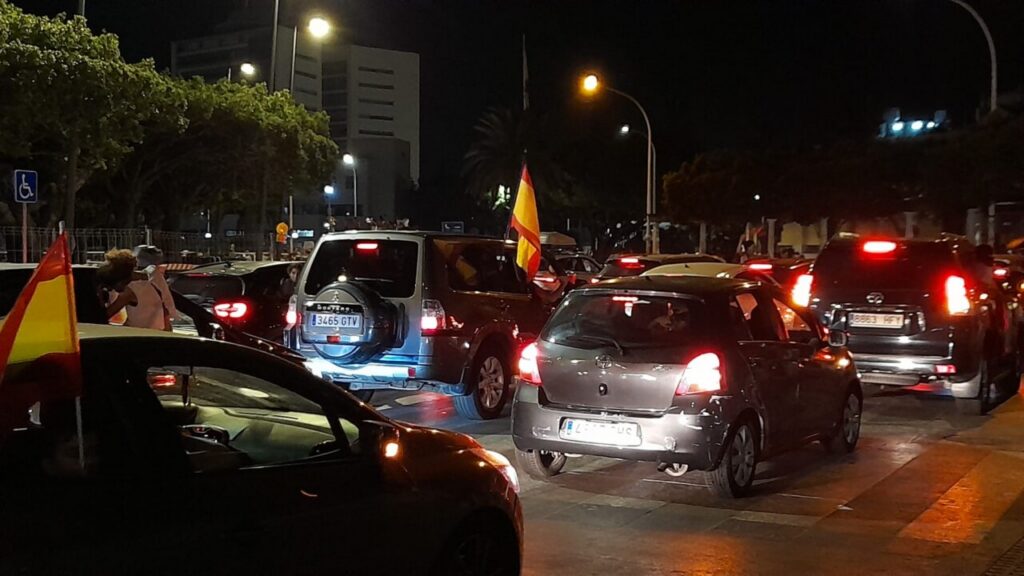 Nighttime street scene with a line of cars in a traffic jam, several red-yellow-red flags visible from car windows indicating a parade or protest.