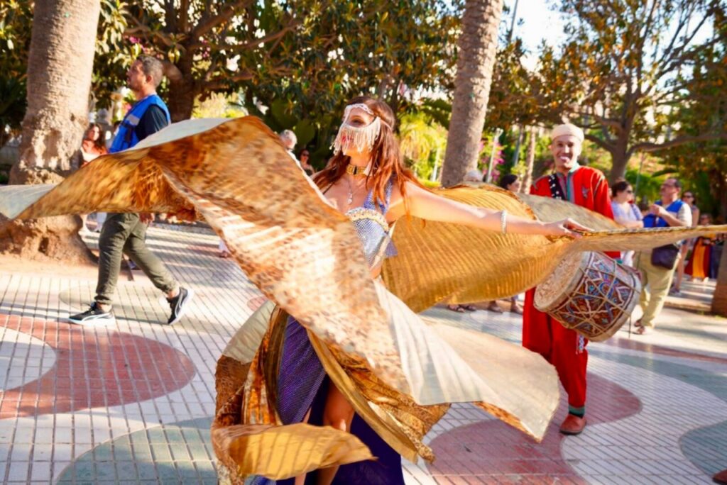 Dancer in a shimmering gold costume with large flowing wings performing in a sunny park with onlookers nearby.