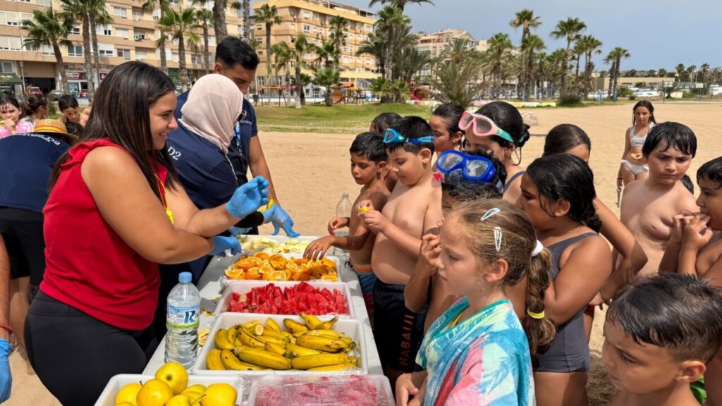 Children at a sunny beach line up to get fruit from a volunteer table with sliced oranges, watermelon, and bananas nearby.