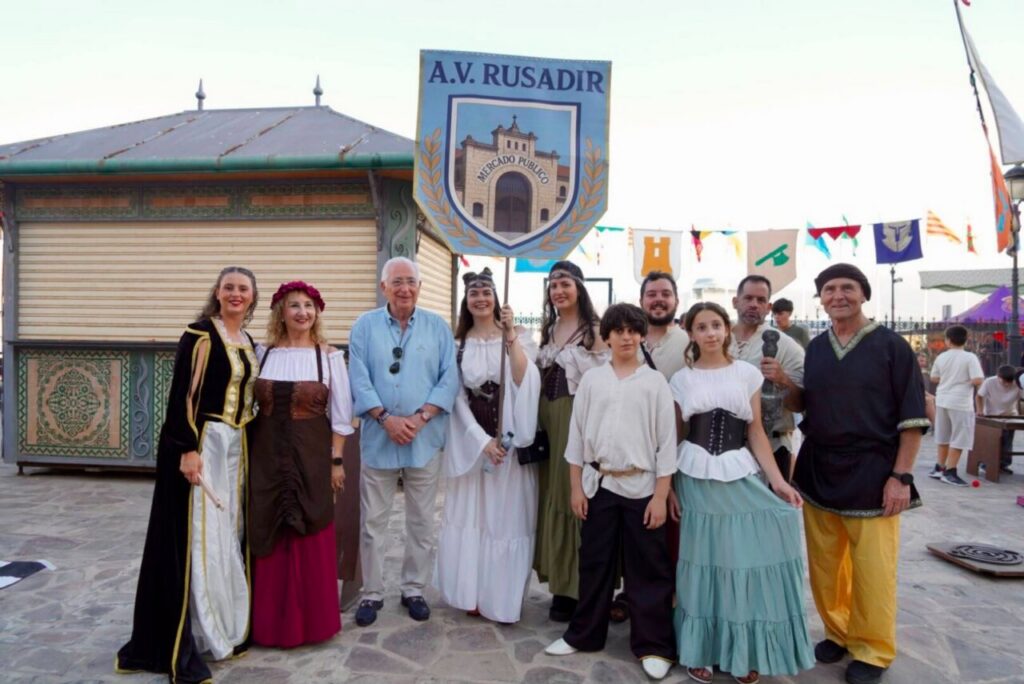 Group of people in medieval costumes posing together at an outdoor festival, a shield banner reading A.V. Rusadir overhead behind them.