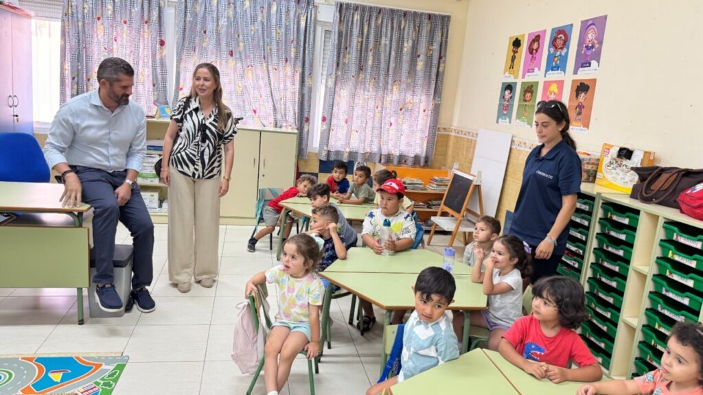Two adults stand at the front of a bright classroom as several young children sit at green tables listening.