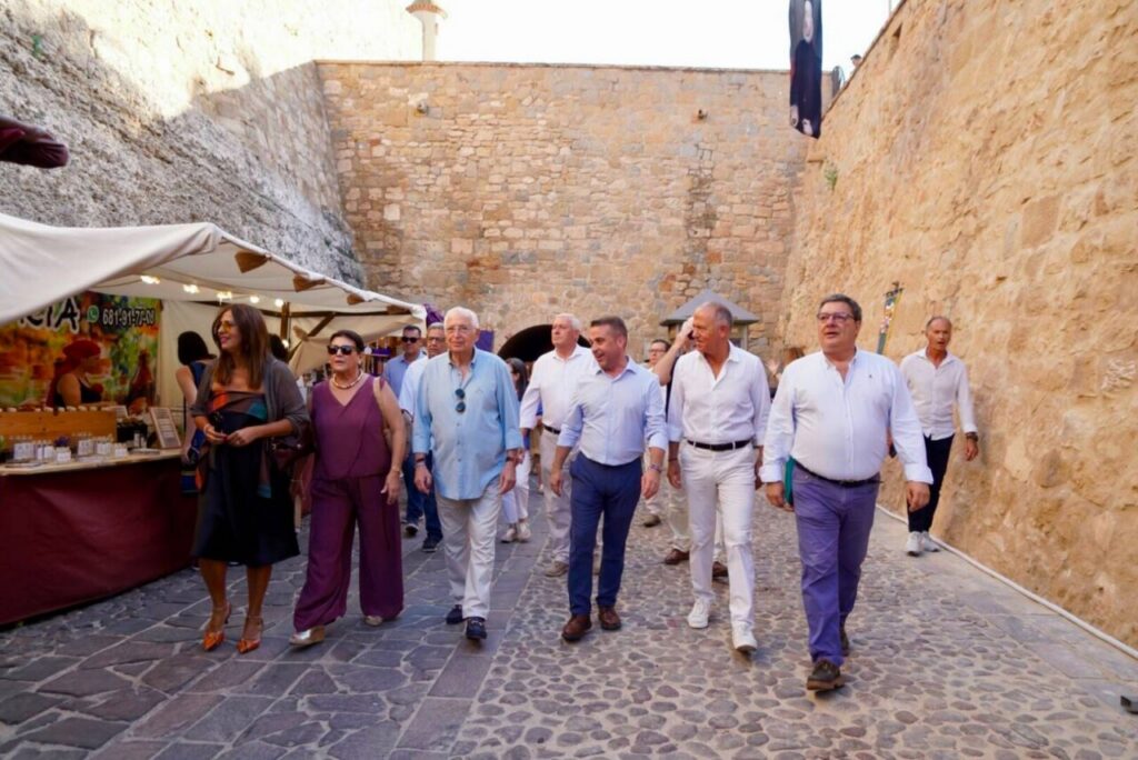 Group of casually dressed adults walking along a cobblestone street beside market stalls in a historic stone-walled area on a sunny day.