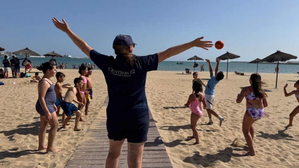 Coach with arms raised directing a beach game while children chase an orange ball on a sunny sandy beach.