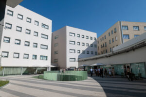 Modern courtyard between white and beige buildings, with a green circular railing and groups of people under umbrellas under a blue sky.