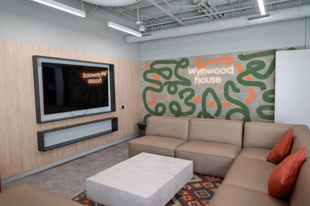 Modern lounge area with a beige sectional sofa and orange cushions, a concrete coffee table, and a colorful mural reading 'wynwood house' on a brick wall behind.