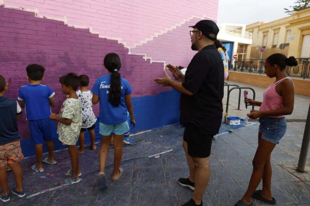 Children line up along a pink brick wall as they paint a mural in purple and pink, guided by an adult in a black shirt and cap.