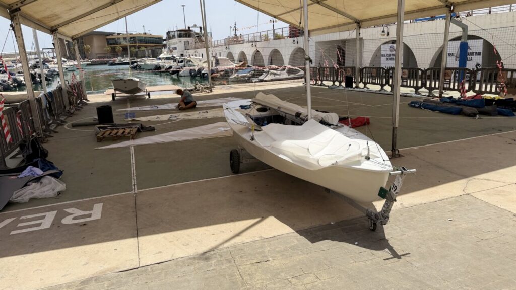 White small sailboat on a trailer under a canopy at a marina, with tools and a person kneeling nearby on the ground level.