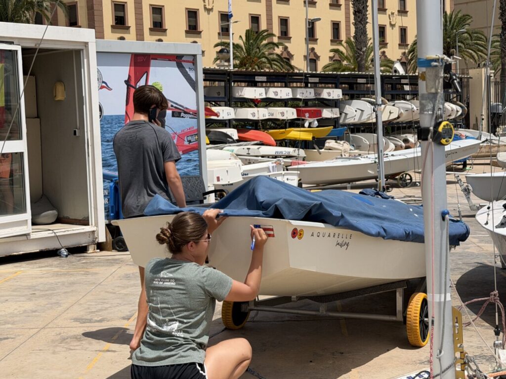 Two people prepare a small sailboat on a wheeled trailer in a marina, one kneeling and signing the hull that reads 'Aquarelle sailing'.