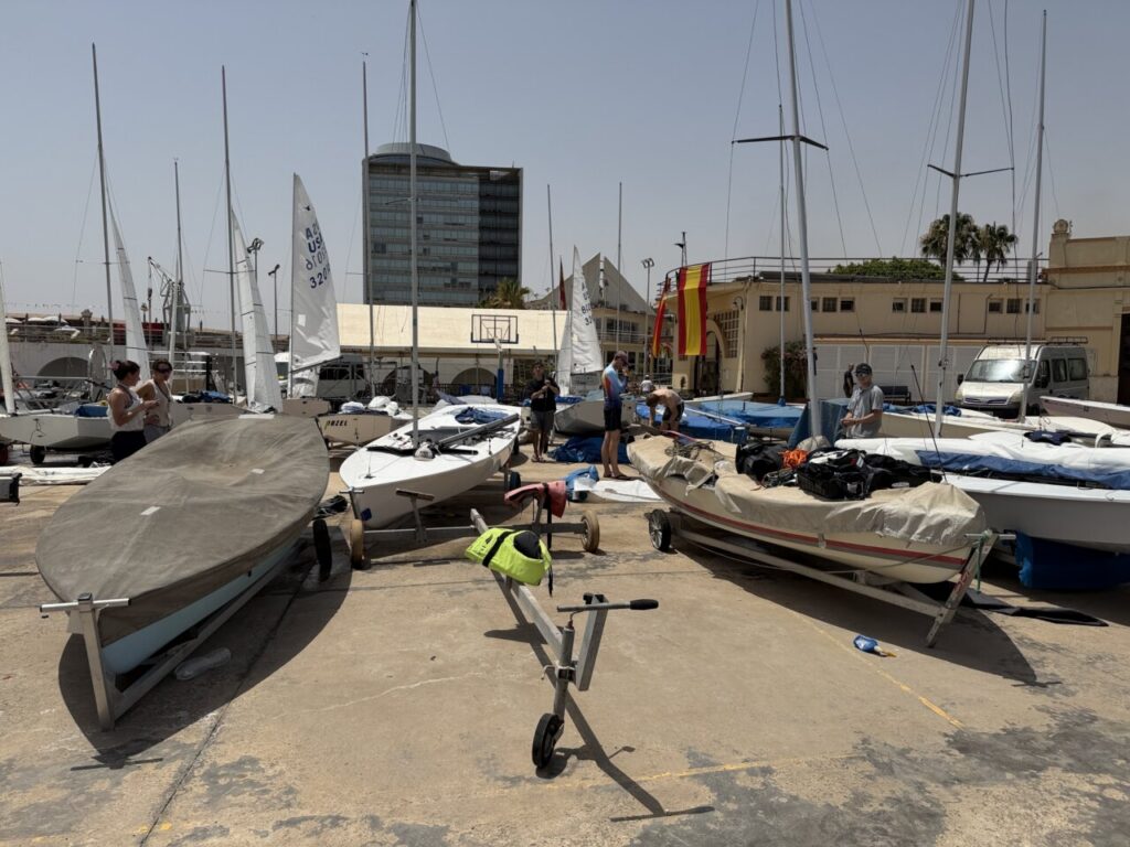 Sailboats on trailers in a sunny marina with people organizing gear and preparing boats on shore.