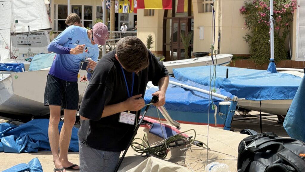 Two men at a marina inspecting and repairing equipment near covered sailboats on a sunny dock.