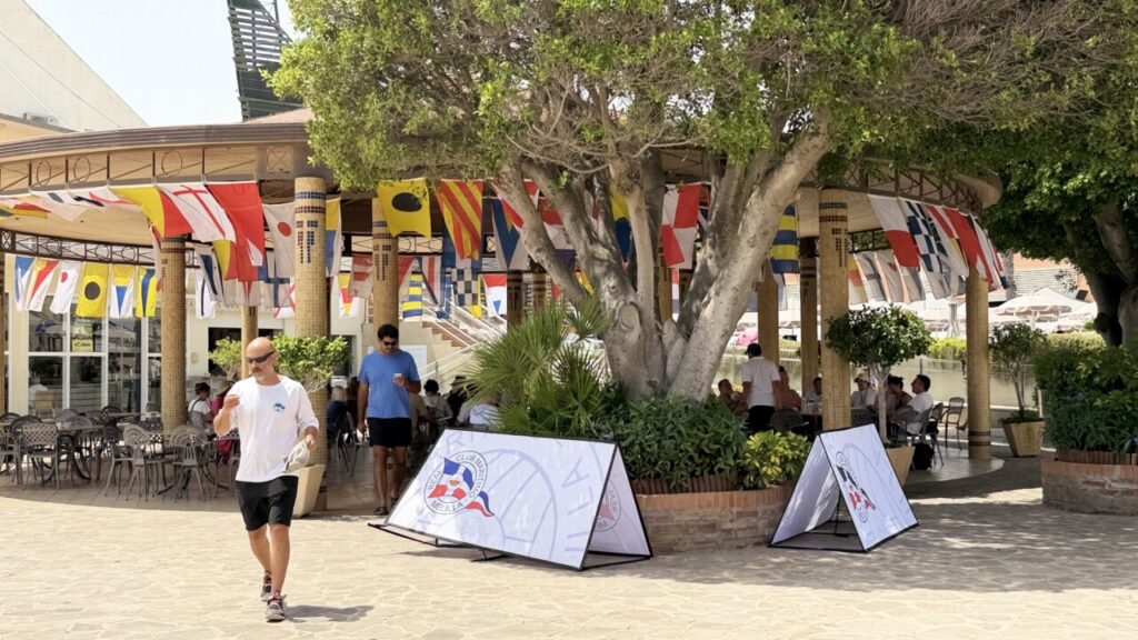 Man in white shirt and black shorts walks past an outdoor cafe beneath colorful nautical flags; a large tree and wicker tables in the background.