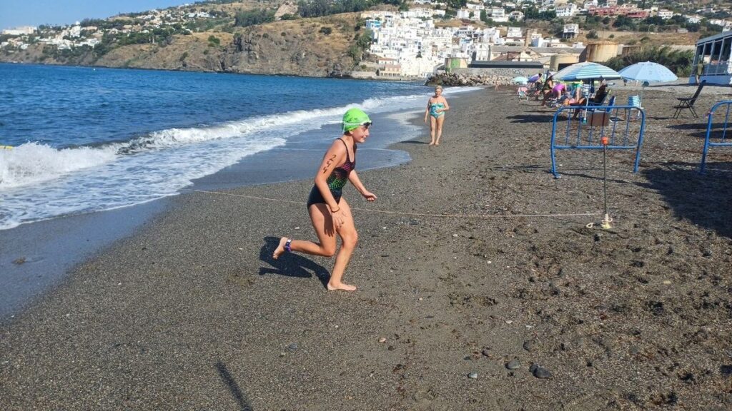 Swimmer in a green cap and black swimsuit runs along a pebbly beach as waves roll in, with people and umbrellas in the background.