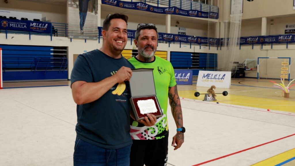 Two men in a gym pose, one presenting a box with a plaque as an award and smiling for the camera.