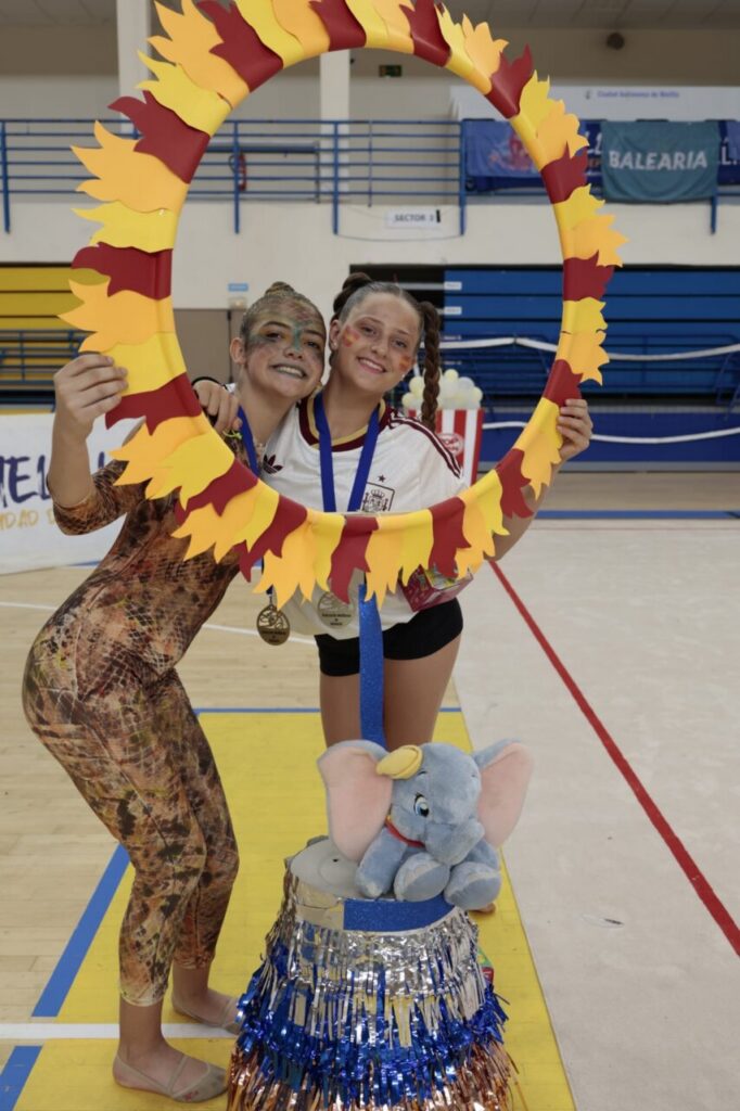 Two smiling gymnasts pose together inside a gym, holding a large yellow‑red leaf wreath and wearing medals.
