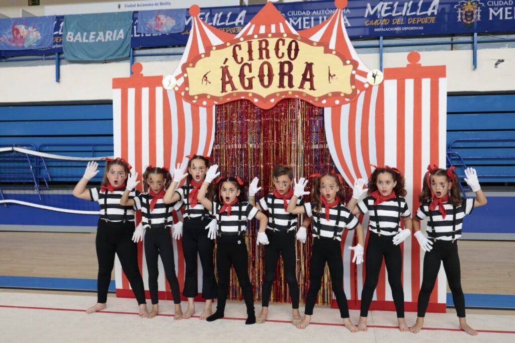 Group of children in mime-style costumes (black-and-white stripes, red scarves, white gloves) posing in front of a red-and-white striped circus backdrop with a sign reading Circo Agora in a gymnasium setting.