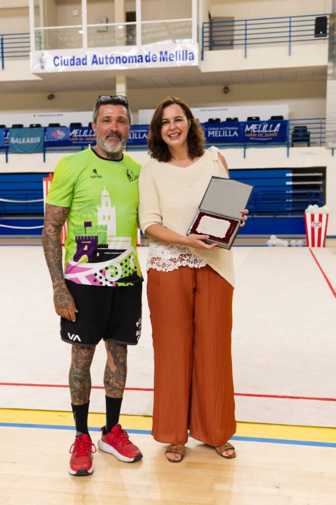 Two people stand in a gym, a tattooed man in a neon shirt next to a smiling woman holding an open award plaque, with Melilla banners in the background.