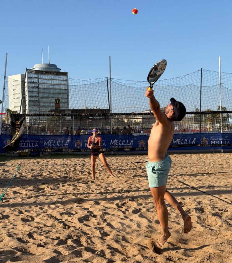Man jumping to strike a beach tennis ball with a paddle over the net on a sandy court; woman player watches from the background on a sunny day.
