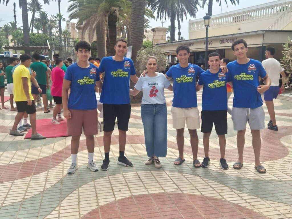 Six youths pose for a group photo in a sunny plaza with palm trees; five wear blue T-shirts that read 'Los Cachorros Del Monte' and one girl in a white T-shirt with a red cherry graphic in the center.