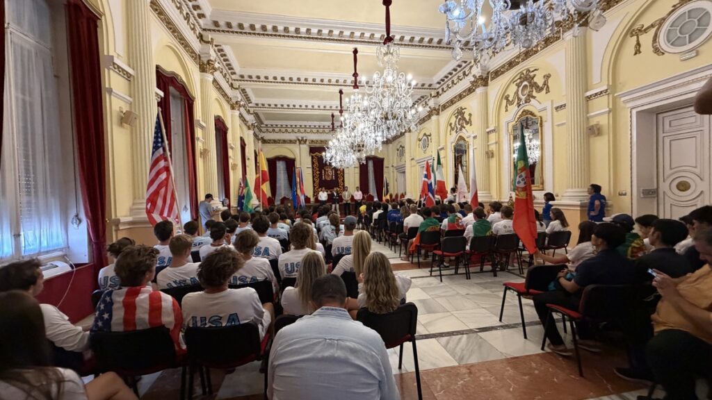 Grand ornate hall filled with people in USA shirts watching a stage presentation; flags line the walls.