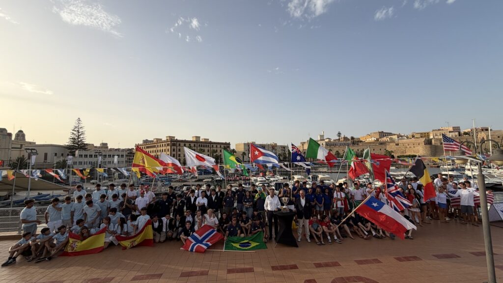 Large group of people posing with international flags on a waterfront promenade, boats in the background at a marina.