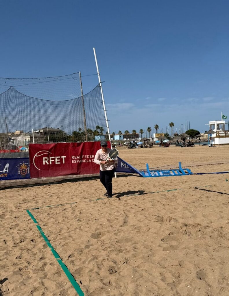 Person walking on a sandy beach tennis court holding a paddle, with a red RFET banner and net nearby on a sunny day.
