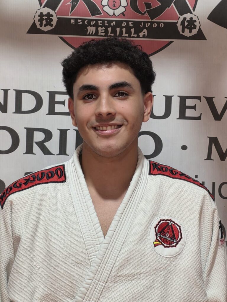 Young man in a white judo gi with red piping and a circular club logo on the chest, smiling at the camera in front of a banner.
