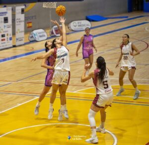 Four female basketball players contest for the ball beneath the hoop on a bright yellow indoor court; two wear white jerseys (numbers 7 and 5) and two wear pink, with others watching.