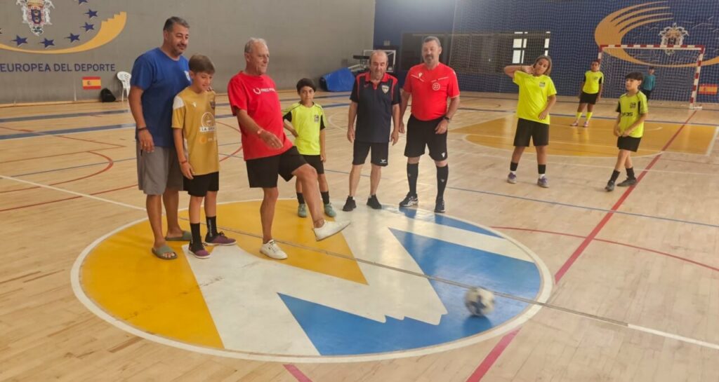 A mixed-age group of players on an indoor futsal court, with a ball rolling near the center circle during a drill or practice session.