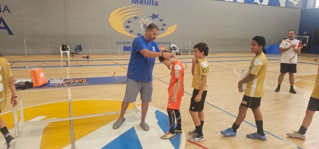 Coach awards a medal to a young player in an orange jersey while teammates line up in a gym gymnasium.