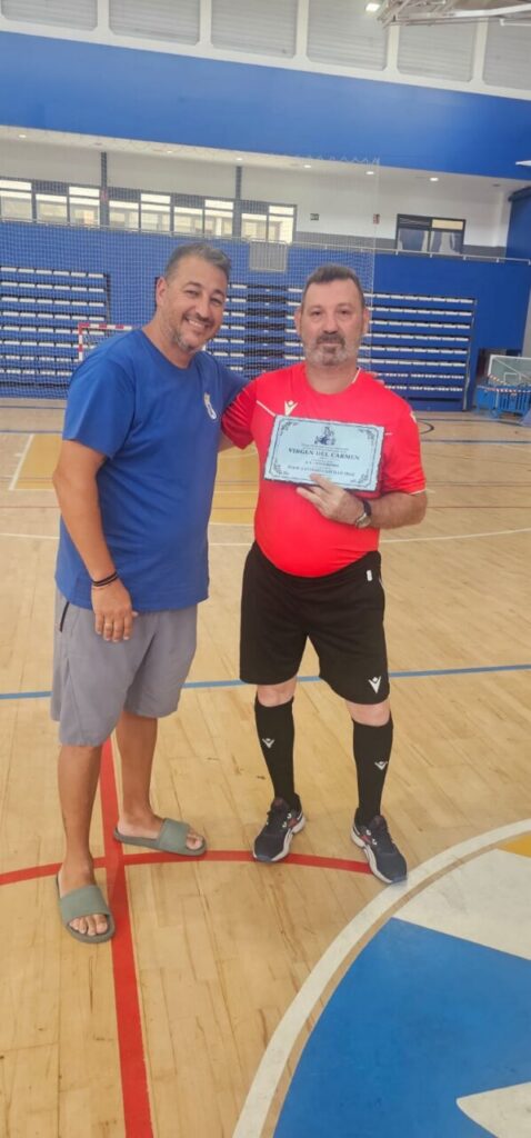 Two men on a gym court; one in blue shirt, the other in red holding a framed award and smiling.