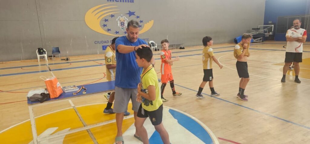Coach adjusts a young player's stance during an indoor youth training session; other kids wait in line on the gym floor.