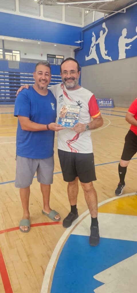 Two men stand in a gym, smiling, as one hands the other a colorful flyer or program.