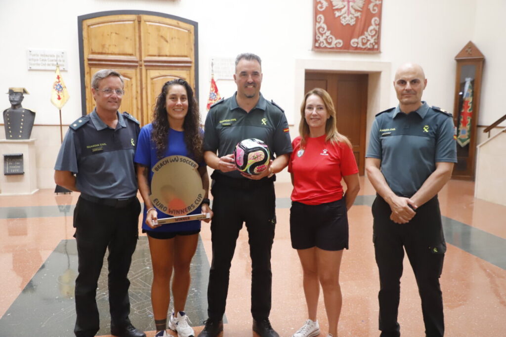 Five people pose in a hallway, three in Guardia Civil uniforms and two in sports attire, holding a round trophy and a soccer ball image.
