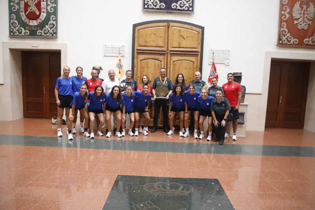 Team of athletes in blue shirts posing with officials in a ceremonial hall; center figure holds a shield, with flags in the background.