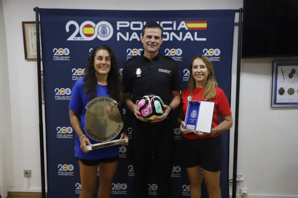 Three people posing with awards at a Policía Nacional event backdrop; left woman holds a circular 'Euro Winners Cup' trophy, center man holds a colorful soccer ball, right woman has a plaque.