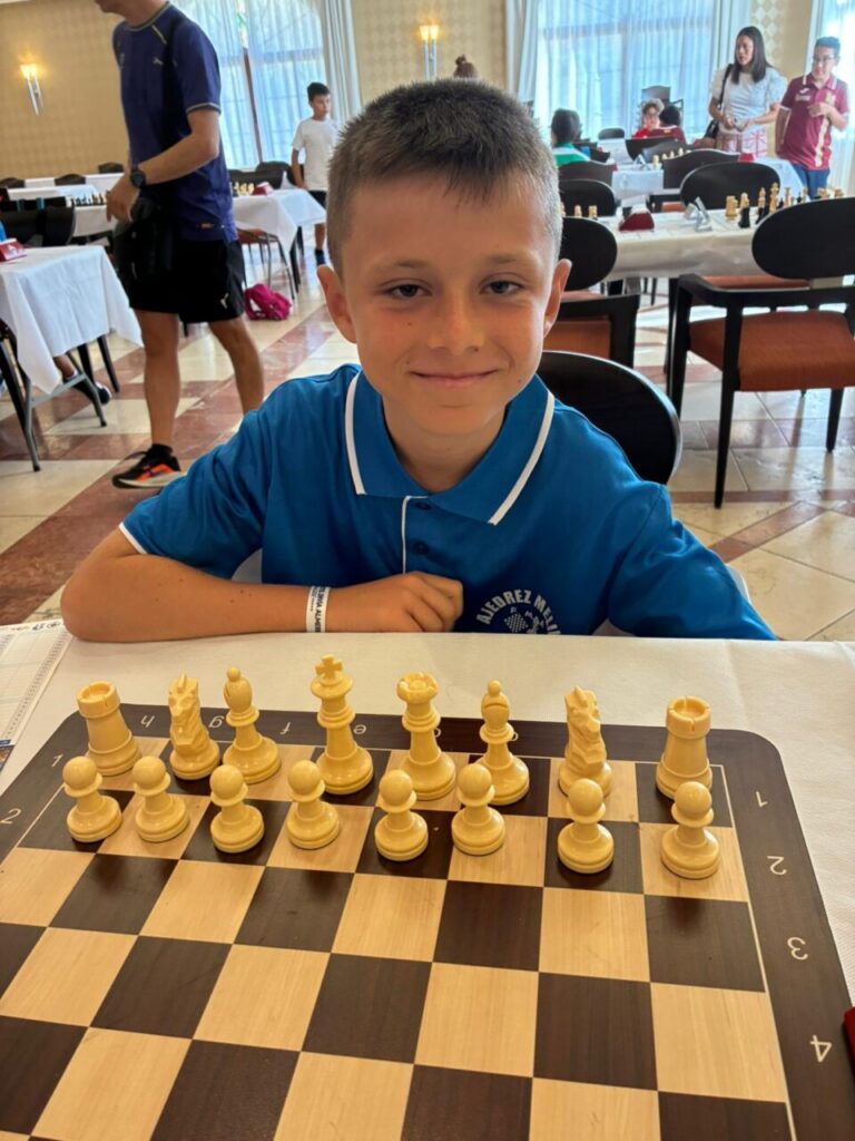 Smiling young boy in a blue polo at a chess tournament, seated at a table with a chessboard and pieces, with players and onlookers in the background.