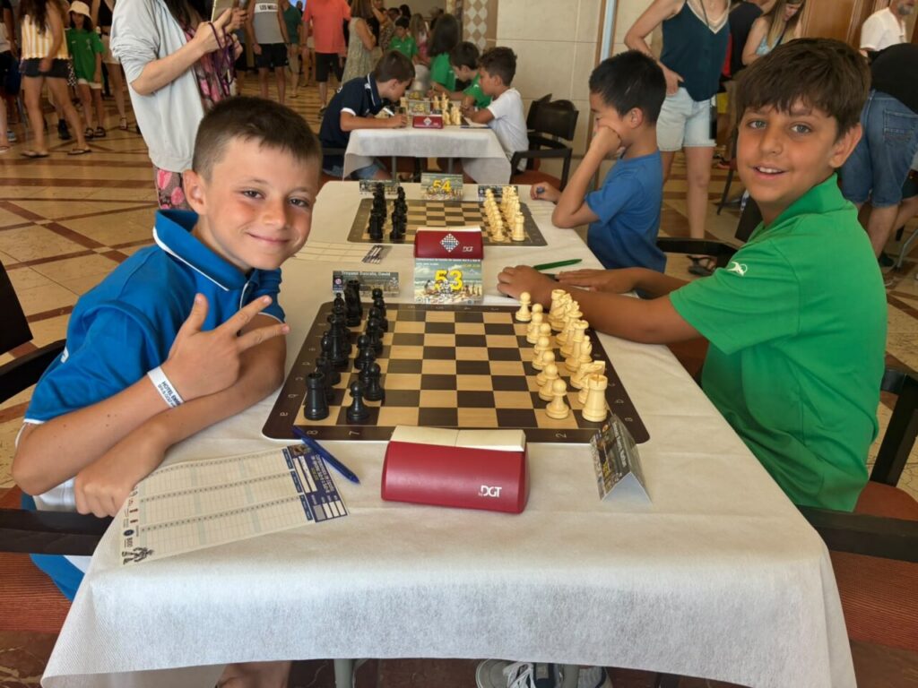 Two boys sit at a chessboard during a kids' tournament, smiling at the camera with other players in the background.