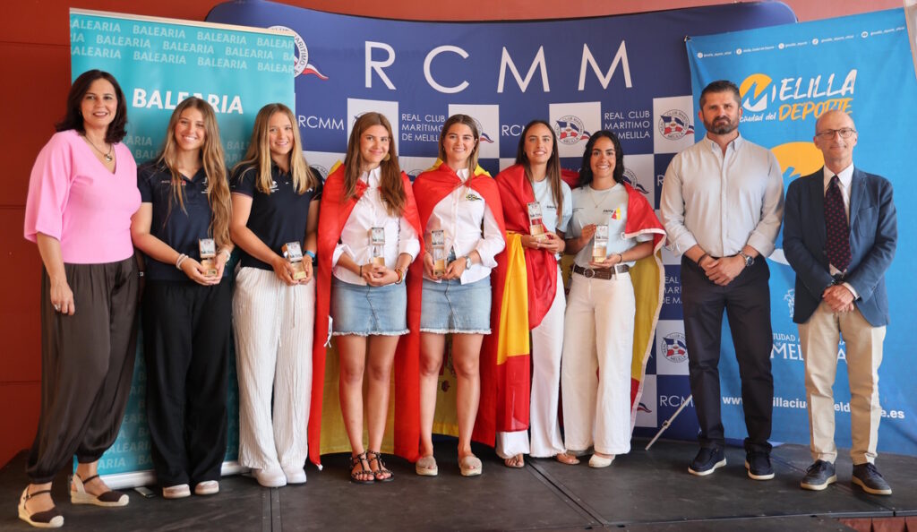 Group of young women standing on a stage, each holding a trophy, with two men and a woman in pink on either side, in front of banners reading RCMM and Balearia; some wear red capes like flags, suggesting an award ceremony.