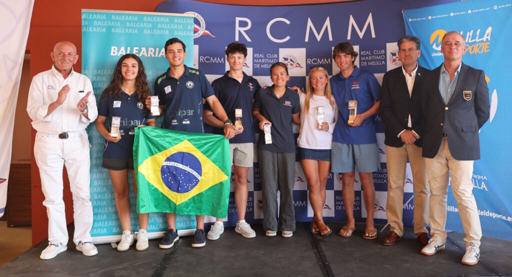 Group of eight young athletes on a stage, holding trophies and a Brazilian flag, flanked by officials, at a sports awards ceremony.