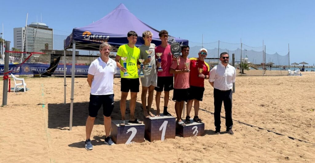 Group of men on a beach podium holding trophies, winners 1st to 3rd place under a purple canopy by the sea.