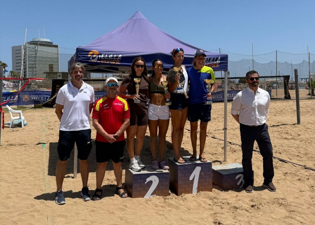 Five beach volleyball players on a three-step podium hold trophies, flanked by two officials on a sunny sandy court with banners nearby.