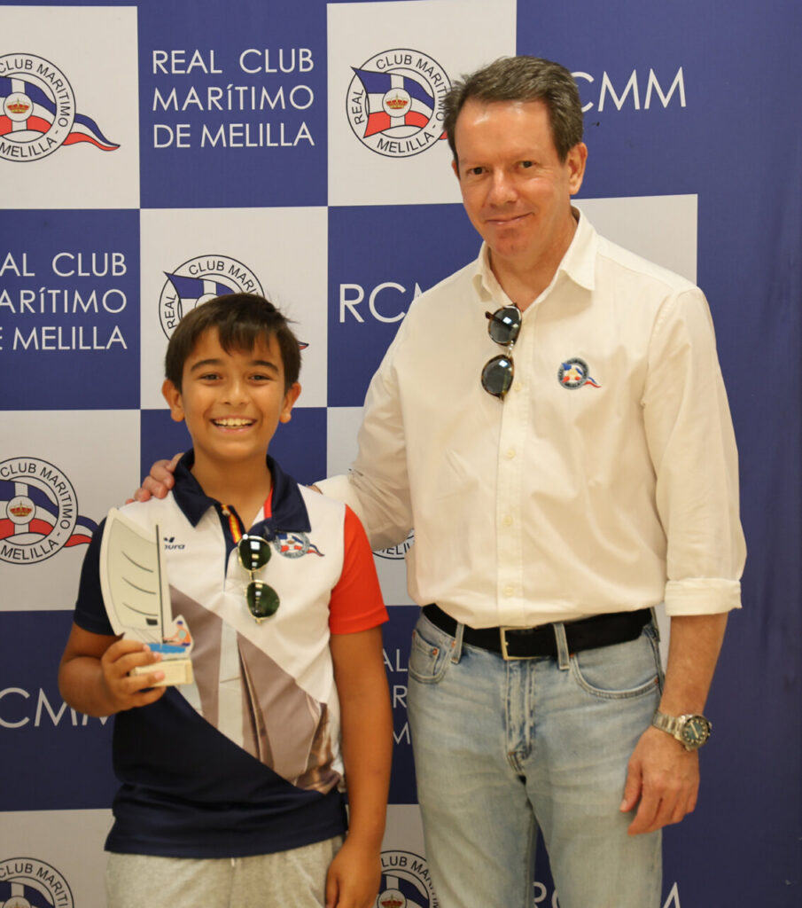 Smiling boy in a polo shirt holds a sailing trophy, posing with a man in a white shirt in front of a blue-and-white club banner.