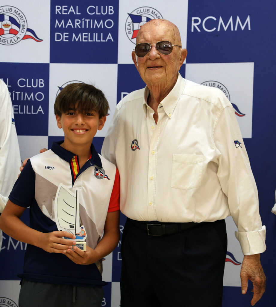 Boy and an older man posing together at a sailing club awards backdrop; the boy holds a trophy.