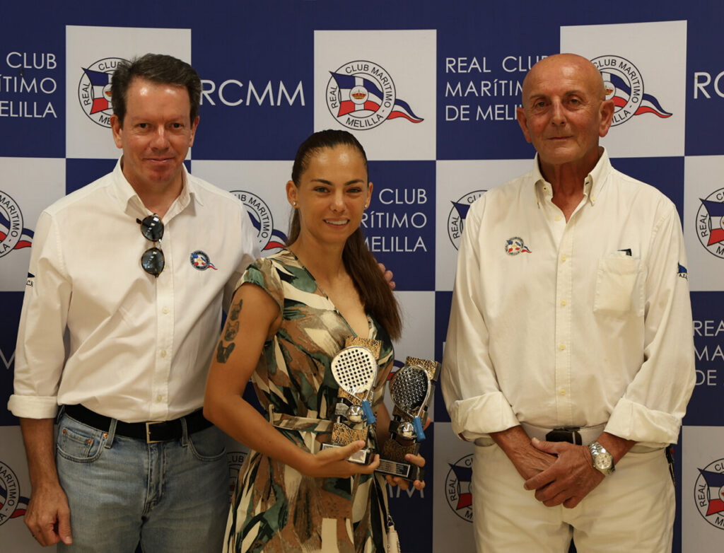 Three people posing at a Real Club Marítimo event; the woman in the center holds two trophies, flanked by two men in white shirts with a logo backdrop.