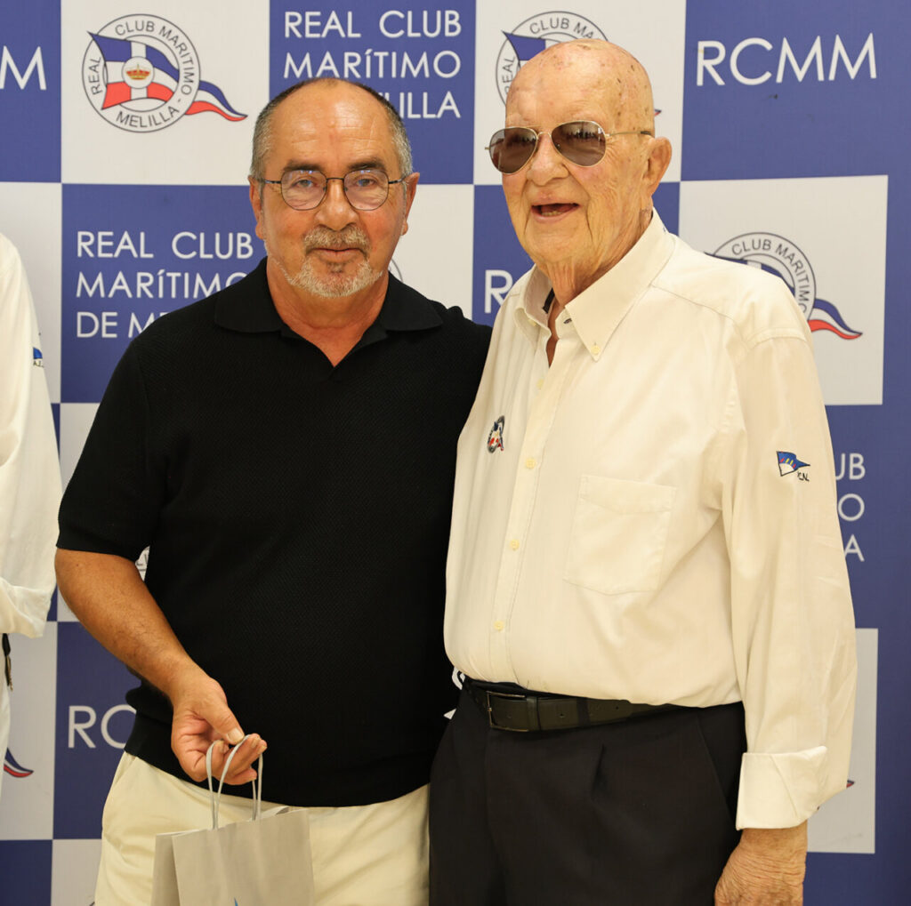Two men pose for a photo in front of a Real Club Marítimo backdrop; one wears a black polo, the other a white shirt with sunglasses, holding gift bags.