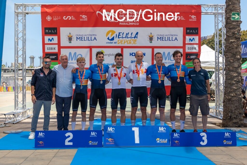 Nine cyclists and staff pose on a blue winner’s podium with medals, in front of a red event banner and sponsor logos.