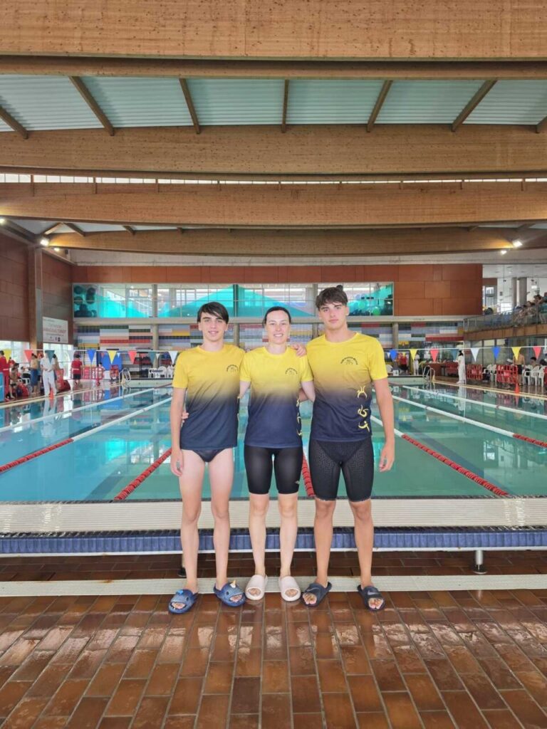 Three swimmers pose together at an indoor pool, wearing matching gradient yellow-to-navy shirts and shorts, standing on the pool deck.