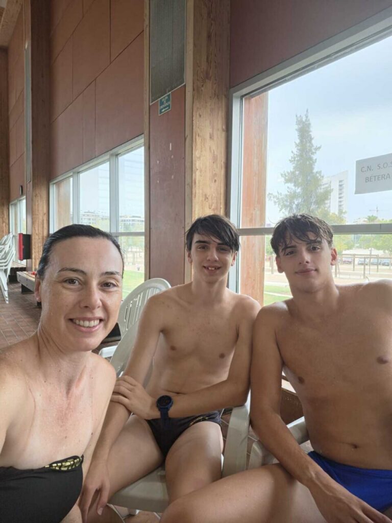 Three people in swimwear sit on plastic chairs inside a pool area, smiling at the camera.